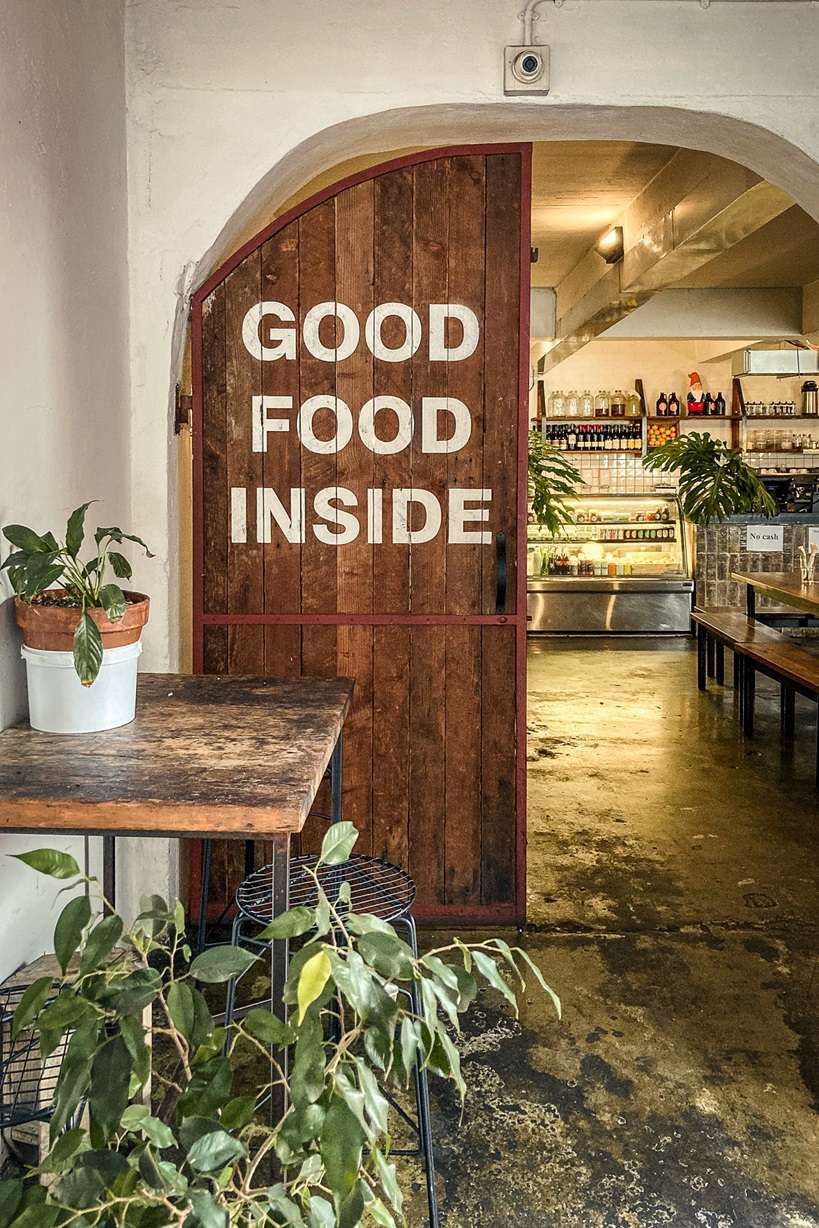 Rustic restaurant entrance with Good Food Inside painted on wooden door surrounded by green plants